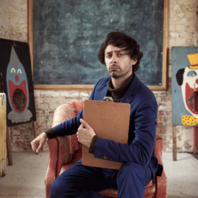 French comedian Marcel Lucont sits on a brown chair wearing a blue suit and holding a clipboard, with a serious expression on his face.There is a picture of a clown in the background.