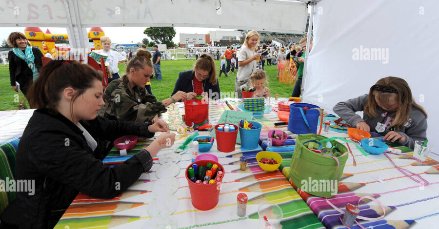 A group of people sat around a table under a white marquee doing arts and crafts in a park.