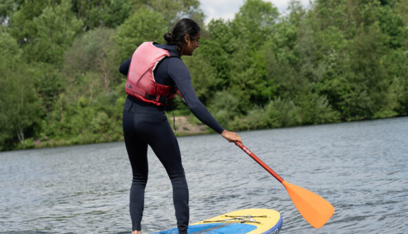 A young person wearing a wet suit and life jacket paddle boaring on a lake.