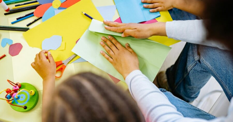 Children and an adult working on a colorful craft project with paper, scissors, and other art supplies on a yellow table.