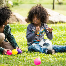 Two young people sit on a grassy lawn outside, playing with brightly coloured small plastic balls.