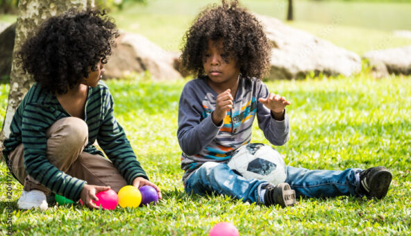 Two young people sit on a grassy lawn outside, playing with brightly coloured small plastic balls.