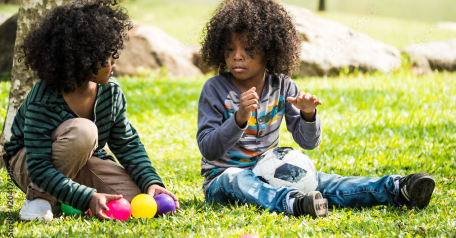 Two young people sit on a grassy lawn outside, playing with brightly coloured small plastic balls.