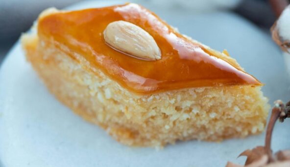A close-up of a slice of basbousa, a Middle Eastern semolina cake, on a white plate. The basbousa is topped with a single almond.