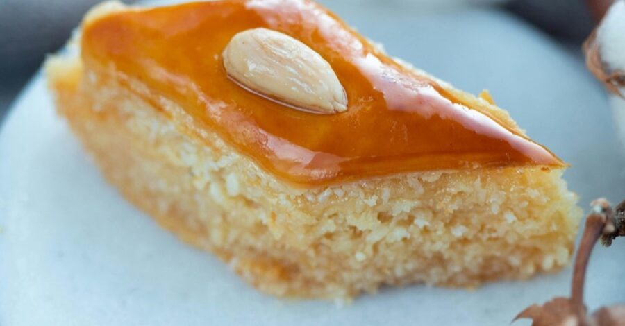 A close-up of a slice of basbousa, a Middle Eastern semolina cake, on a white plate. The basbousa is topped with a single almond.