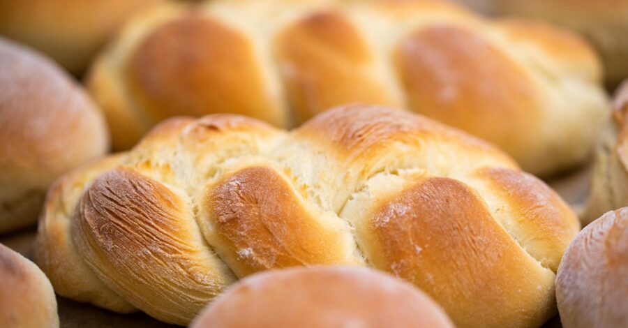 A close-up image of freshly baked challah bread. The bread is golden-brown, braided, and has a light, fluffy texture. Multiple loaves are visible in the background, all exhibiting a warm, inviting appearance with a slight dusting of flour.