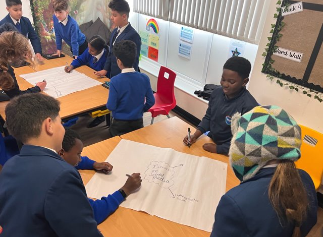 A group of children dressed in school uniforms sat on chair around a table. There is a large piece of white paper which they are writing on.