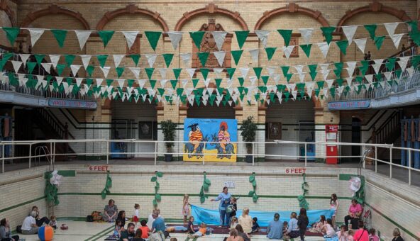 people are sitting on colourful blankets and cushions in the Gala Pool at Victoria baths. there is green and white bunting hanging from the ceiling.