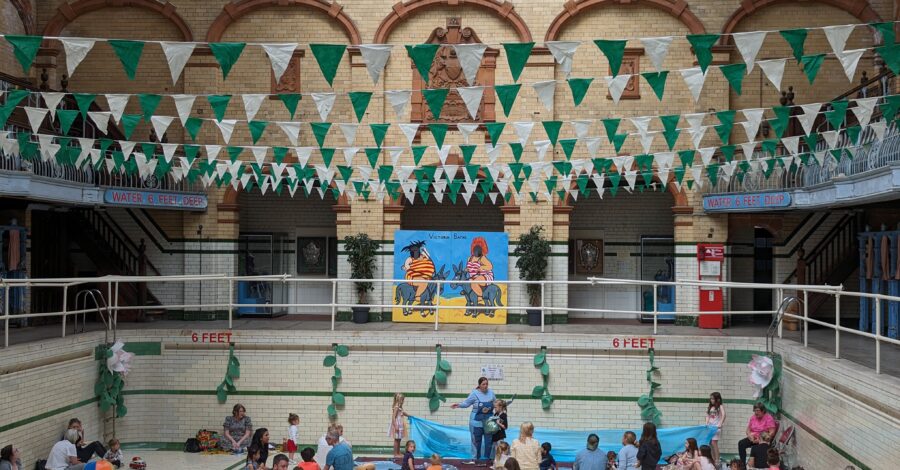 people are sitting on colourful blankets and cushions in the Gala Pool at Victoria baths. there is green and white bunting hanging from the ceiling.