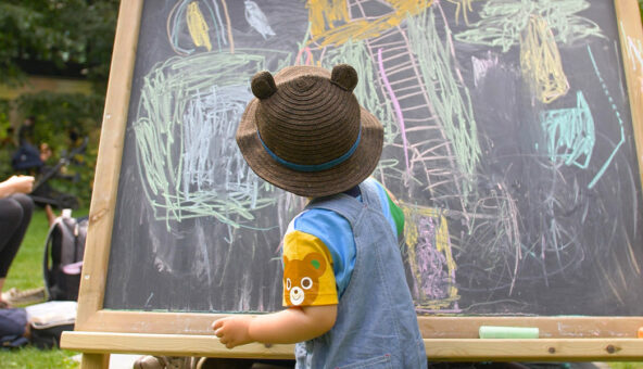 A child, wearing a brown hat with bear ears on it, making marks with coloured chalk on a chalkboard.