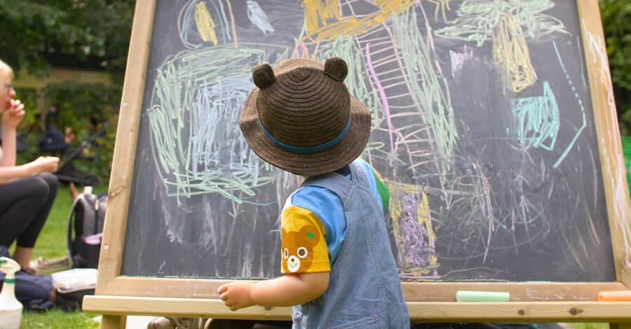 A child, wearing a brown hat with bear ears on it, making marks with coloured chalk on a chalkboard.