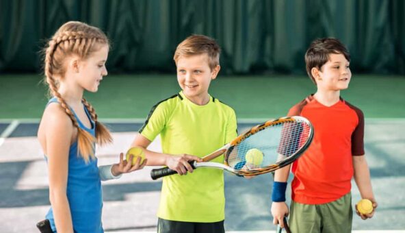 Three children stand holding a tennis racket and a ball ready to play tennis.