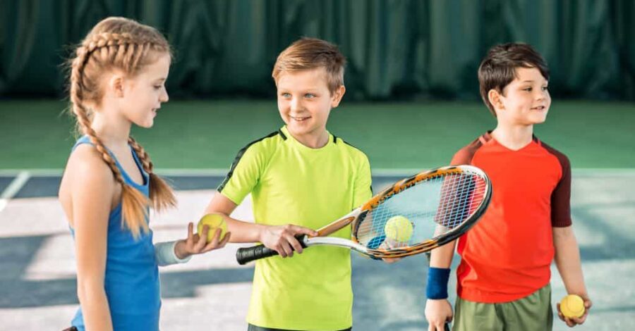 Three children stand holding a tennis racket and a ball ready to play tennis.