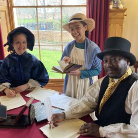 A family dressed in period costume sat at a table writing and smiling.