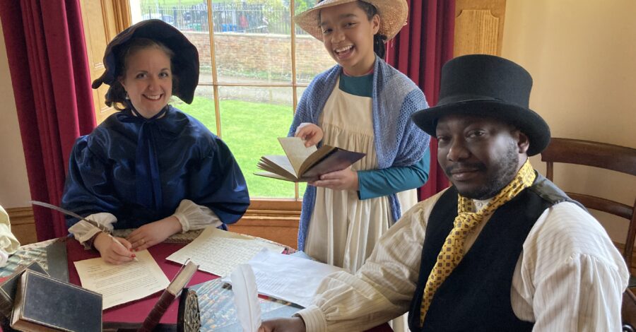 A family dressed in period costume sat at a table writing and smiling.