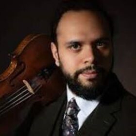 The head and shoulders of violinist and musician Faz Shah. He wears a white shirt, tie and jacket and holds a violin on his shoulder.