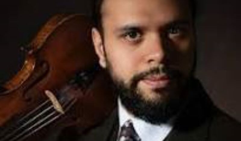 The head and shoulders of violinist and musician Faz Shah. He wears a white shirt, tie and jacket and holds a violin on his shoulder.