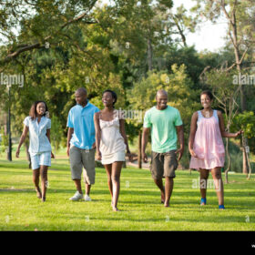 Five people walk on grass through a park. They are dressed as if it is a warm day with summer clothes like dresses, shorts and t-shirts.