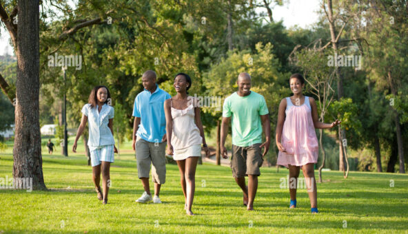 Five people walk on grass through a park. They are dressed as if it is a warm day with summer clothes like dresses, shorts and t-shirts.
