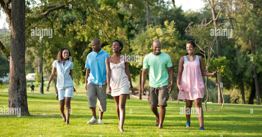 Five people walk on grass through a park. They are dressed as if it is a warm day with summer clothes like dresses, shorts and t-shirts.