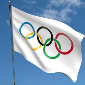 The Olympic flag, consisting of five rings of blue, yellow, black, green and red on a white background, blowing in a breeze under a blue sky.