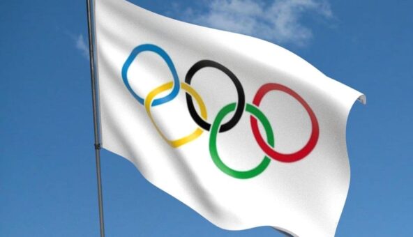The Olympic flag, consisting of five rings of blue, yellow, black, green and red on a white background, blowing in a breeze under a blue sky.