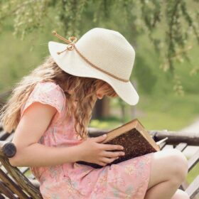 A child, wearing a pink dress and a straw hat, sitting on a chair reading a book outside.