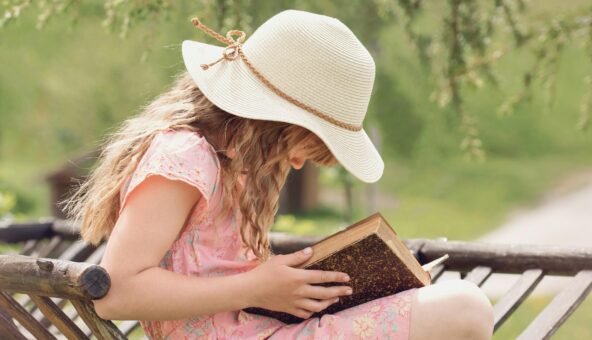 A child, wearing a pink dress and a straw hat, sitting on a chair reading a book outside.