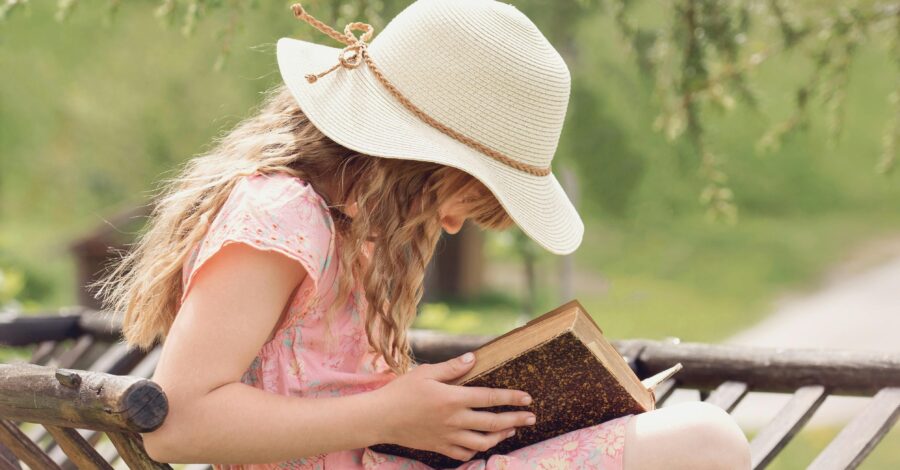 A child, wearing a pink dress and a straw hat, sitting on a chair reading a book outside.