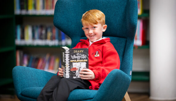 A boy, wearing a red sweatshirt, sits smiling on a blue wing-backed armchair holding a Diary of a Wimpy Kid book.