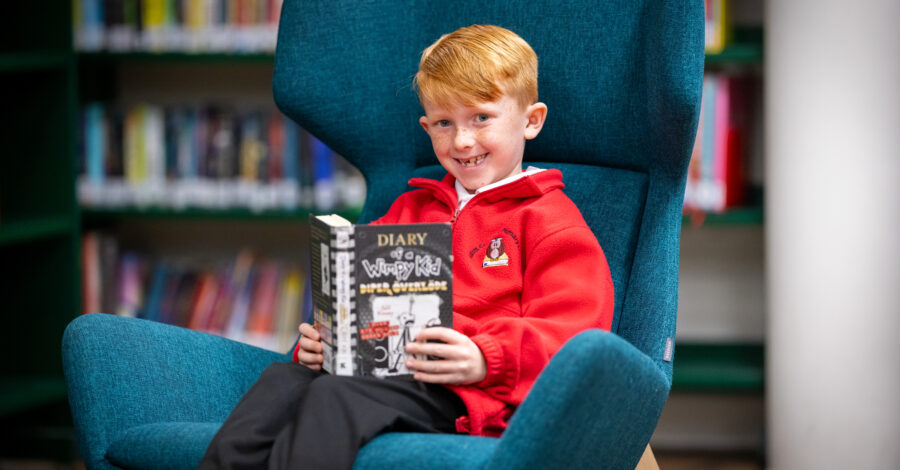 A boy, wearing a red sweatshirt, sits smiling on a blue wing-backed armchair holding a Diary of a Wimpy Kid book.