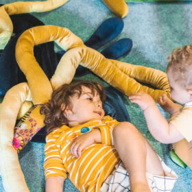 Two babies, one lying down, one sitting up, playing with velvet snake-like soft long objects.