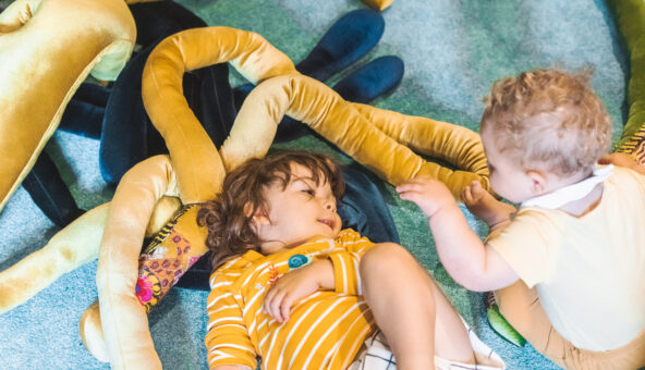 Two babies, one lying down, one sitting up, playing with velvet snake-like soft long objects.