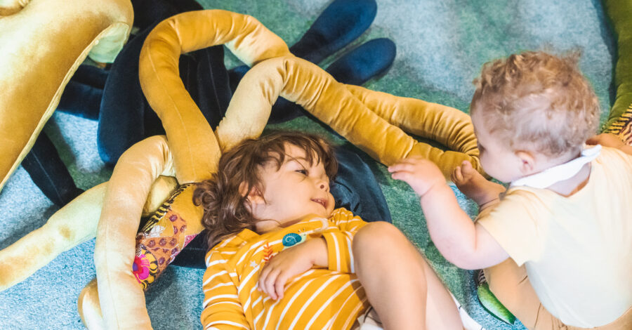 Two babies, one lying down, one sitting up, playing with velvet snake-like soft long objects.