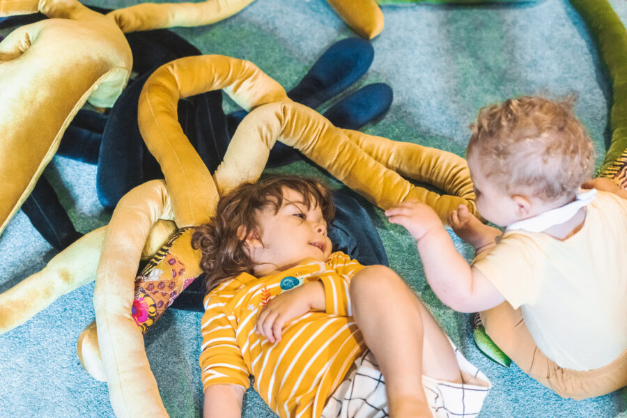 Two babies, one lying down, one sitting up, playing with velvet snake-like soft long objects.