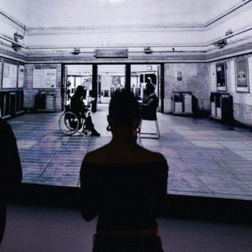 An audience members watches on as two people, one sat on a chair and one in a wheelchair, have a conversation.