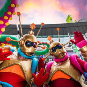 Two figures dressed up as aliens with gold helmets and masks on , orange antenas and gold, orange and red bodysuits. They are waving at the camera.