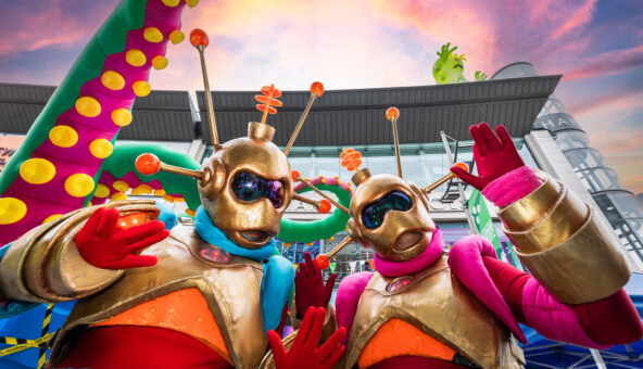 Two figures dressed up as aliens with gold helmets and masks on , orange antenas and gold, orange and red bodysuits. They are waving at the camera.