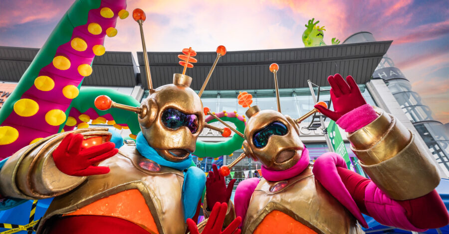 Two figures dressed up as aliens with gold helmets and masks on , orange antenas and gold, orange and red bodysuits. They are waving at the camera.