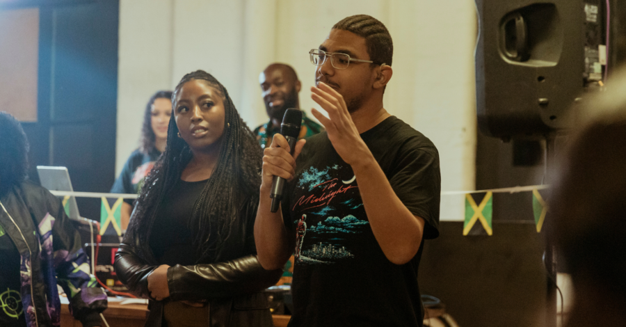 A young man stands talking into a microphone. Several young people watch him.