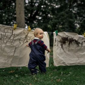 Toddler standing on grass with a paintbrush in hand turning to the right to look at the camera. He is wearing a navy blue rainsuit with a burgundy and navy blue long sleeved tiop underneath. He is standing in front of large cream coloured sheets that have been strung up on a washing line. The sheets have mud marks making on.