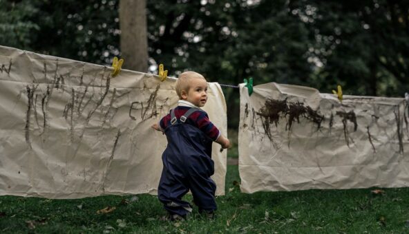 Toddler standing on grass with a paintbrush in hand turning to the right to look at the camera. He is wearing a navy blue rainsuit with a burgundy and navy blue long sleeved tiop underneath. He is standing in front of large cream coloured sheets that have been strung up on a washing line. The sheets have mud marks making on.