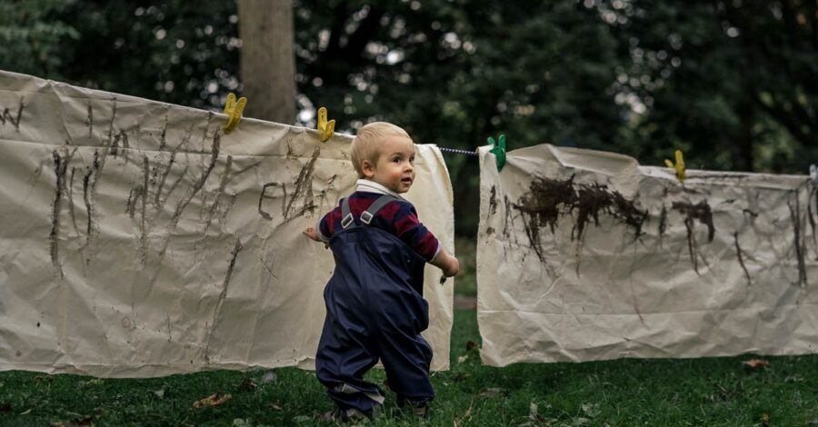 Toddler standing on grass with a paintbrush in hand turning to the right to look at the camera. He is wearing a navy blue rainsuit with a burgundy and navy blue long sleeved tiop underneath. He is standing in front of large cream coloured sheets that have been strung up on a washing line. The sheets have mud marks making on.