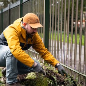 A man wearing a yellow jacket and cap planting near a metal fence.