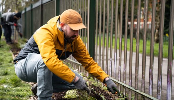 A man wearing a yellow jacket and cap planting near a metal fence.