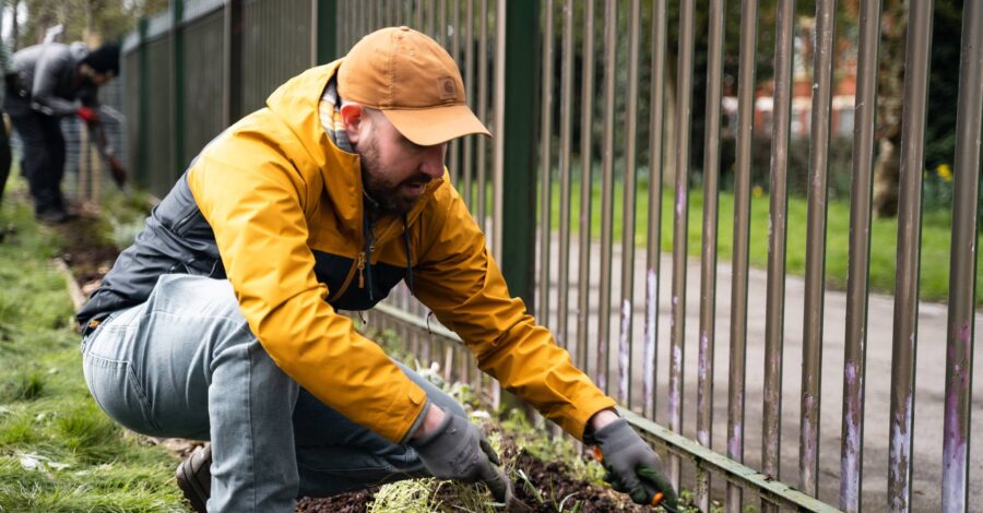 A man wearing a yellow jacket and cap planting near a metal fence.