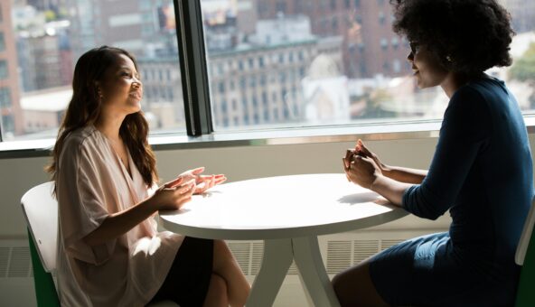Two people sat on chairs at a white table chatting.