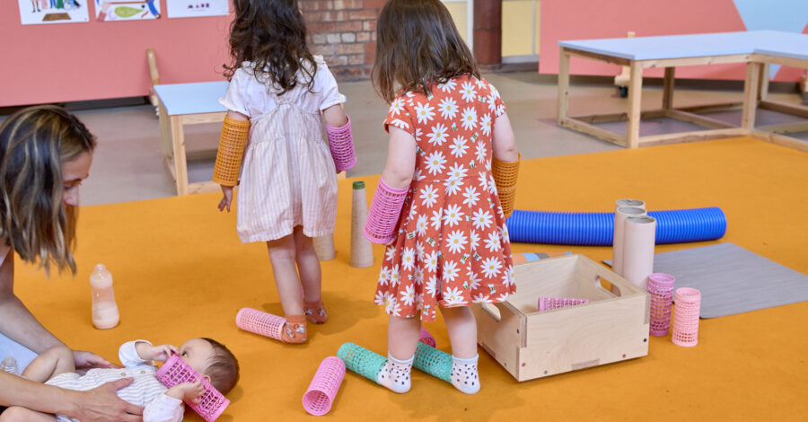 Two pre-school ages children walking with bobbins on their feet and on their arms with their backs to the camera. Parent to the left of shot knelt down looking over a baby lying on an orange rug playing with the same style of bobbin.