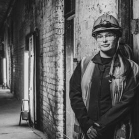 Black & White image of a young women wearing hi-viz clothes and a hard hat standing by a rough brick wall