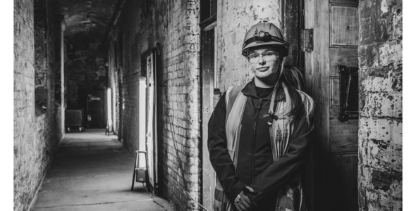 Black & White image of a young women wearing hi-viz clothes and a hard hat standing by a rough brick wall